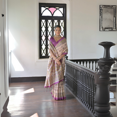 Woman wearing a beige silk saree with intricate floral motifs and a purple border, accessorized with a choker necklace and bangles, standing in a sunlit vintage interior with wooden flooring, black railing, and a window with stained glass panels.