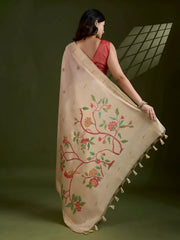  Woman wearing an elegant beige silk saree featuring delicate floral motifs and a hand-painted pallu, paired with a sleeveless red blouse and traditional jewelry, gracefully posing in a modern studio with olive green walls and patterned window light.