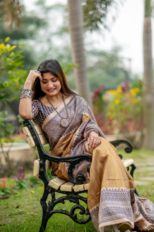 Woman in a beige saree with Madhubani print standing near a pond with lotus flowers in a lush garden setting