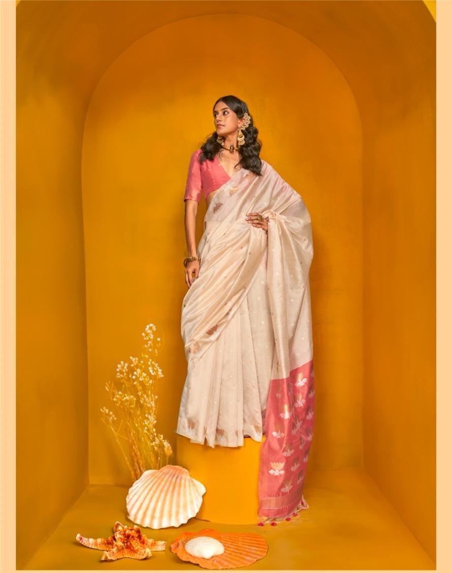 Model in beige zari work saree with pink floral border and matching blouse, styled with gold jewellery and seashell decor in yellow arched backdrop