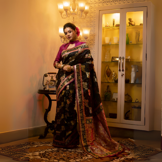 Woman wearing a black Banarasi silk saree with intricate multicolor floral zari work and a magenta blouse, accessorized with gold jewelry, standing in a warmly lit vintage interior featuring a chandelier, antique telephone, and a glass display cabinet.