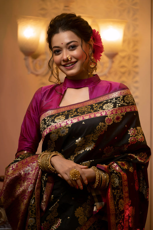 Woman wearing a black Banarasi silk saree with intricate multicolor floral zari work and a magenta blouse, accessorized with gold jewelry, standing in a warmly lit vintage interior featuring a chandelier, antique telephone, and a glass display cabinet.