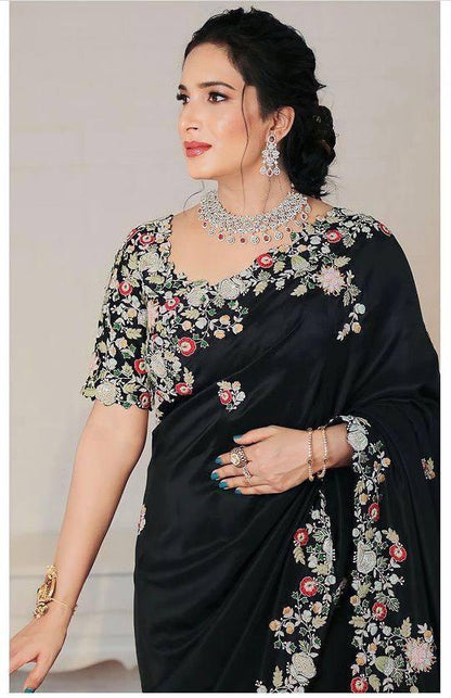 Woman wearing a black saree with intricate embroidery work, accessorized with a necklace and earrings, standing in a room with wooden flooring