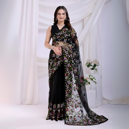  Woman wearing a black net saree with vibrant multicolor floral embroidery and a matching sleeveless blouse, posing in a modern white studio with draped curtains and white floral arrangements in the background.