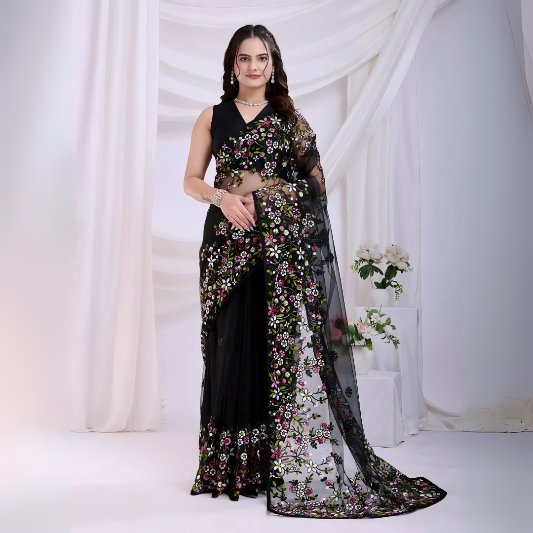  Woman wearing a black net saree with vibrant multicolor floral embroidery and a matching sleeveless blouse, posing in a modern white studio with draped curtains and white floral arrangements in the background.