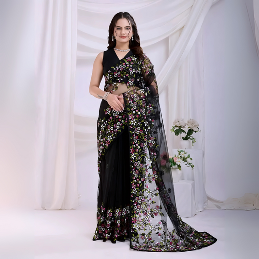  Woman wearing a black net saree with vibrant multicolor floral embroidery and a matching sleeveless blouse, posing in a modern white studio with draped curtains and white floral arrangements in the background.