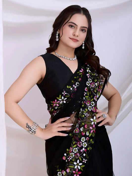  Woman wearing a black net saree with vibrant multicolor floral embroidery and a matching sleeveless blouse, posing in a modern white studio with draped curtains and white floral arrangements in the background.