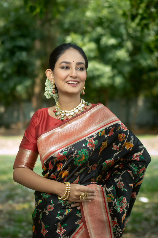 Woman wearing a black silk saree with vibrant multicolor floral motifs and a contrasting coral pink border, paired with a matching blouse and traditional jewelry, elegantly standing in front of a large, ancient tree with exposed roots in a serene outdoor setting.