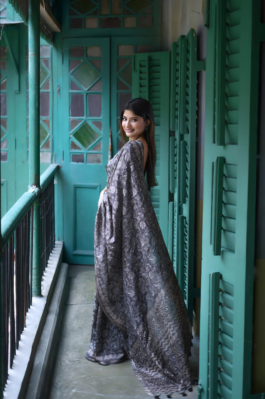 Woman wearing a black and grey floral printed saree with a matching blouse, accessorized with statement rings, standing on a vintage balcony with teal green shutters and doors