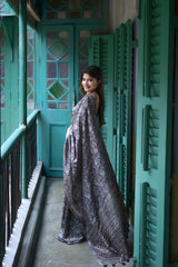 Woman wearing a black and grey floral printed saree with a matching blouse, accessorized with statement rings, standing on a vintage balcony with teal green shutters and doors