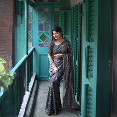 Woman wearing a black and grey floral printed saree with a matching blouse, accessorized with statement rings, standing on a vintage balcony with teal green shutters and doors.