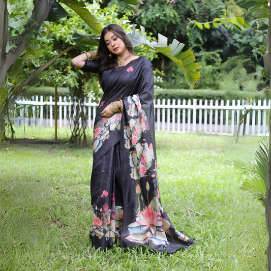Woman wearing a black saree with large handpainted lotus and leaf floral motifs, accessorized with traditional jewelry, posing gracefully outdoors in a lush green garden with tropical plants and a white picket fence in the background.
