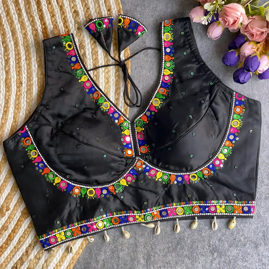 Black blouse featuring vibrant mirror work and traditional Kutch multicolor embroidery along the neckline and hem, adorned with scattered sequin details and shell tassels. The blouse includes a stylish back tie-up with matching embroidered tassels and is displayed flat on a textured surface with a floral bouquet, highlighting its festive ethnic appeal.