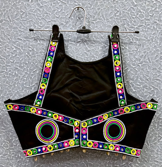 Black blouse featuring vibrant circular and oval mirror work with traditional Kutch multicolor embroidery, adorned with bold geometric borders and shell tassel drops along the hem. Displayed on a hanger against a textured silver background, highlighting its festive and culturally inspired ethnic design.
