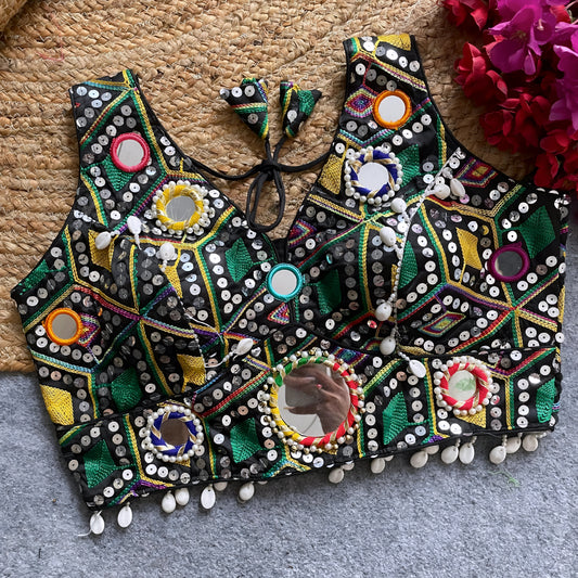Traditional black Gujarati-style blouse richly adorned with vibrant multicolor geometric embroidery and circular mirror work, decorated with colorful thread rings, white cowrie shell trim along the hem, and tasseled tie-back detail, displayed flat on a grey carpet with a woven mat and fuchsia artificial flowers beside it.