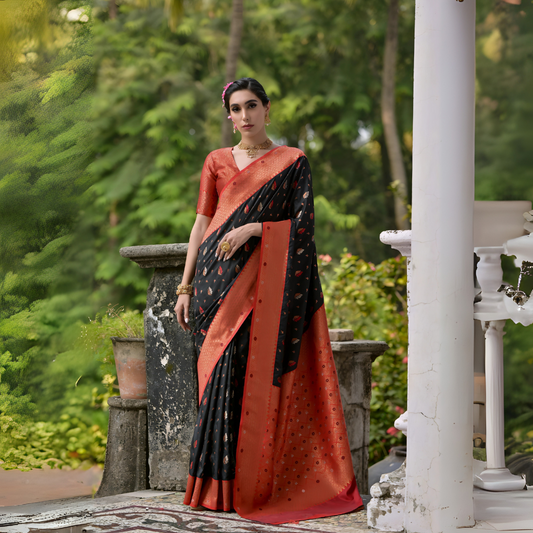  Woman wearing a black and orange silk saree with gold motifs and a contrasting orange blouse, posing outdoors on a veranda with lush green trees in the background.