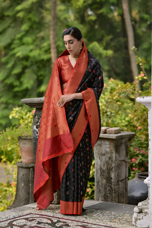  Woman wearing a black and orange silk saree with gold motifs and a contrasting orange blouse, posing outdoors on a veranda with lush green trees in the background.