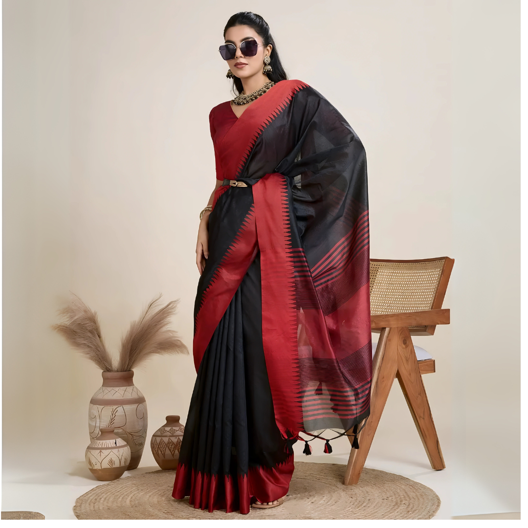 Woman wearing a striking black and red silk saree featuring a bold geometric border and pallu, paired with a matching blouse and statement necklace, elegantly posing beside a wooden chair in a minimalist studio setting with neutral decor, ceramic vases, and pampas grass.
