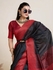 Woman wearing a striking black and red silk saree featuring a bold geometric border and pallu, paired with a matching blouse and statement necklace, elegantly posing beside a wooden chair in a minimalist studio setting with neutral decor, ceramic vases, and pampas grass.