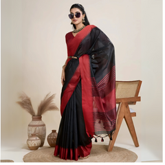 Woman wearing a striking black and red silk saree featuring a bold geometric border and pallu, paired with a matching blouse and statement necklace, elegantly posing beside a wooden chair in a minimalist studio setting with neutral decor, ceramic vases, and pampas grass.