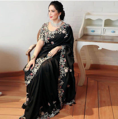 A person wearing a black saree adorned with intricate embroidery work, standing in a room with wooden flooring and light-colored curtains.