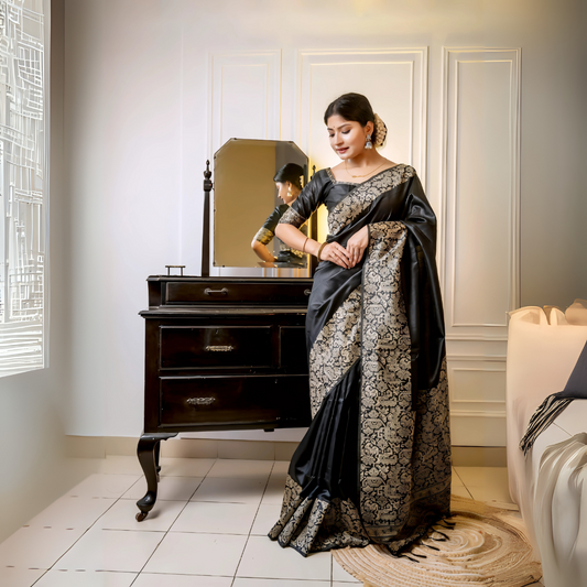 Woman wearing a black silk saree with intricate gold brocade patterns and a matching blouse, standing gracefully beside a vintage black dresser with a mirror in a softly lit studio featuring white paneled walls and elegant decor.
