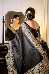 Woman wearing a black silk saree with intricate gold brocade patterns and a matching blouse, standing gracefully beside a vintage black dresser with a mirror in a softly lit studio featuring white paneled walls and elegant decor.