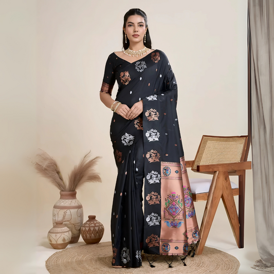Woman wearing a black silk saree adorned with multicolor woven motifs and a contrasting peach pallu featuring intricate designs, paired with a matching blouse and traditional jewelry, elegantly posing beside a wooden chair in a minimalist studio with neutral decor, ceramic vases, and pampas grass.