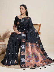 Woman wearing a black silk saree adorned with multicolor woven motifs and a contrasting peach pallu featuring intricate designs, paired with a matching blouse and traditional jewelry, elegantly posing beside a wooden chair in a minimalist studio with neutral decor, ceramic vases, and pampas grass.