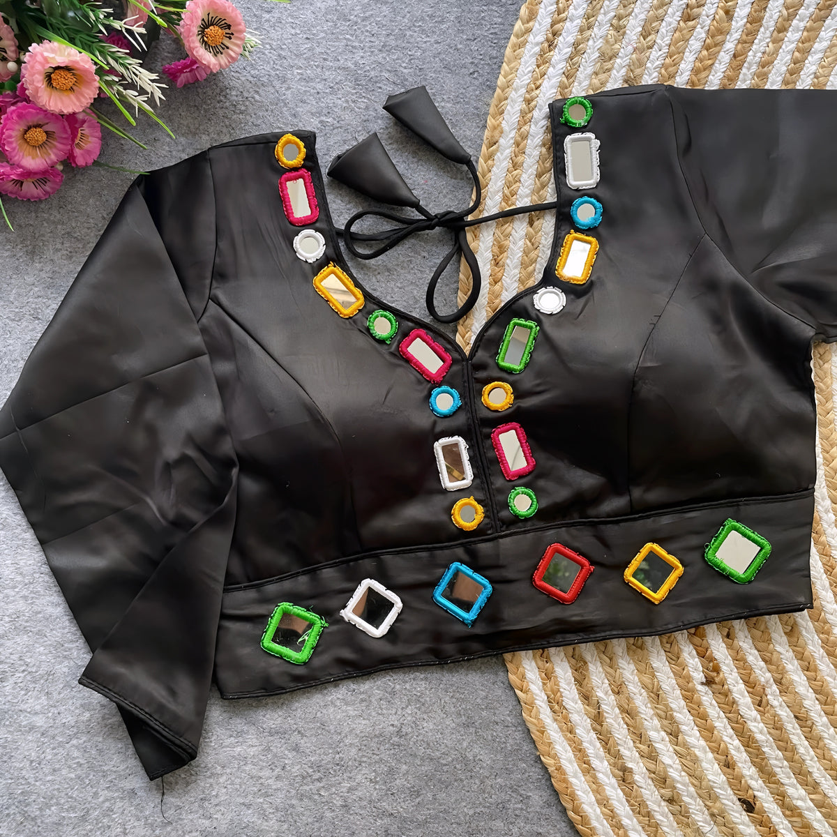 Black blouse decorated with vibrant square and round mirror work featuring multicolored embroidered borders, stylish back tie-up details, and three-quarter sleeves. Displayed flat on a textured grey surface with woven mat and flowers, highlighting its festive and contemporary ethnic appeal.