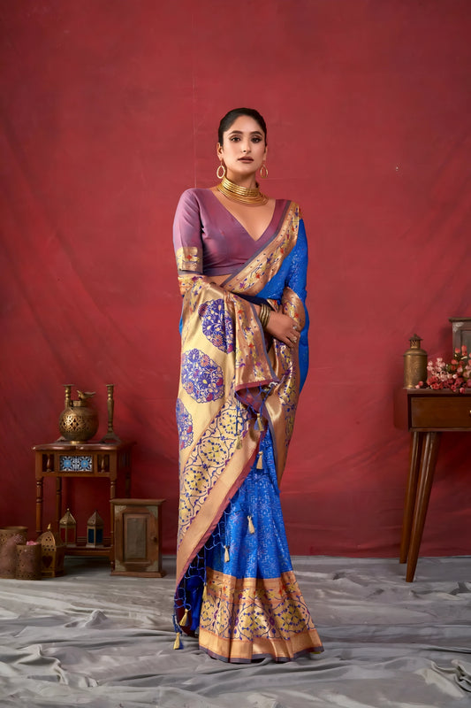 Woman wearing a blue Banarasi silk saree with intricate gold and purple motifs and a gold border, paired with a mauve blouse and traditional gold jewelry, holding a bouquet and standing in front of a deep red vintage-inspired backdrop with classic wooden decor.