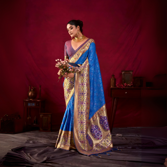 Woman wearing a blue Banarasi silk saree with intricate gold and purple motifs and a gold border, paired with a mauve blouse and traditional gold jewelry, holding a bouquet and standing in front of a deep red vintage-inspired backdrop with classic wooden decor.