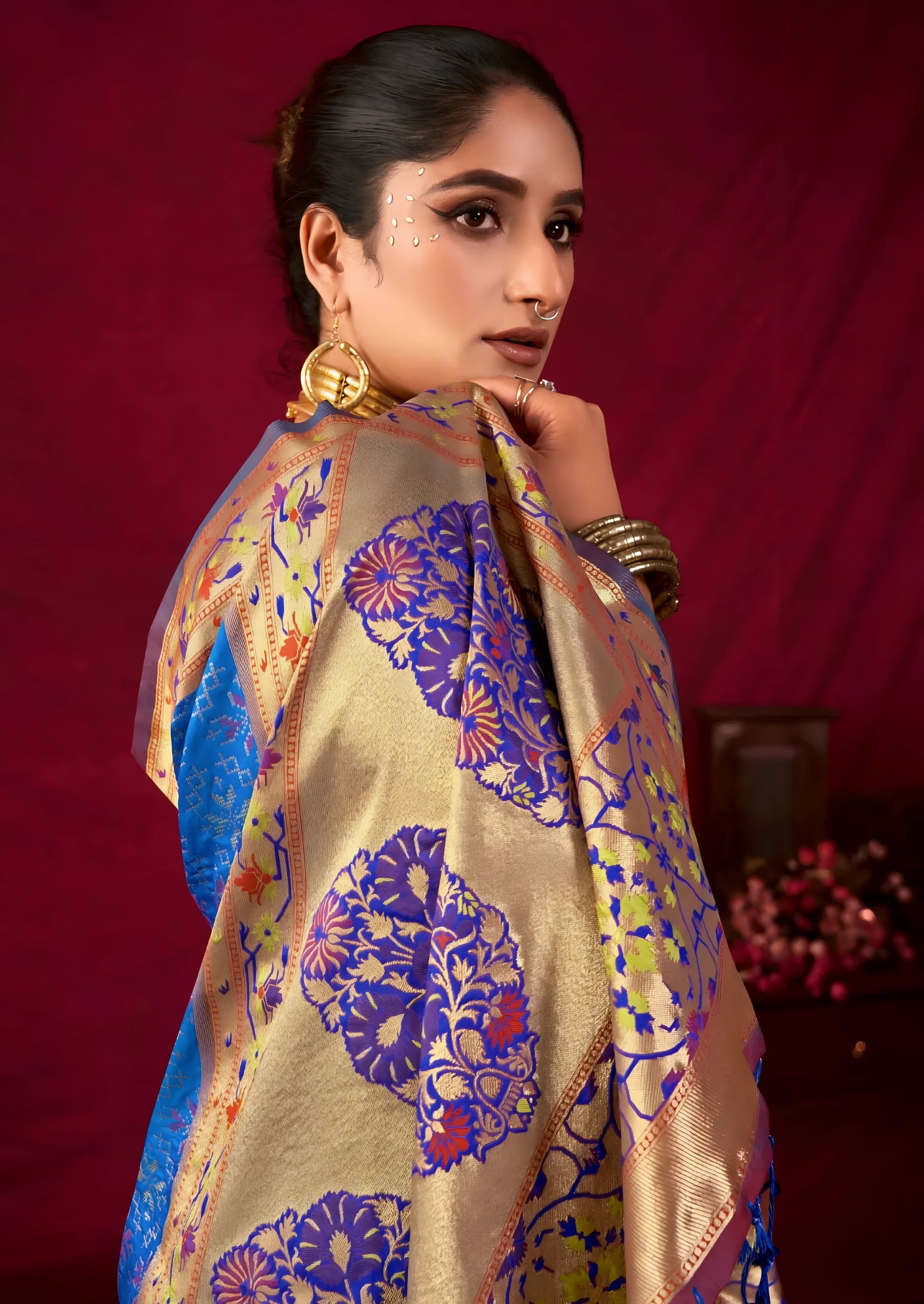 Woman wearing a blue Banarasi silk saree with intricate gold and purple motifs and a gold border, paired with a mauve blouse and traditional gold jewelry, holding a bouquet and standing in front of a deep red vintage-inspired backdrop with classic wooden decor.