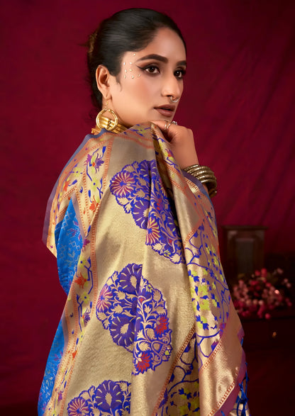 Woman wearing a blue Banarasi silk saree with intricate gold and purple motifs and a gold border, paired with a mauve blouse and traditional gold jewelry, holding a bouquet and standing in front of a deep red vintage-inspired backdrop with classic wooden decor.