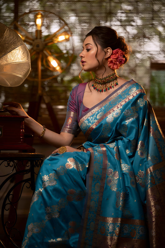 Woman wearing a blue Banarasi silk saree with intricate gold zari motifs and border, paired with a purple blouse and traditional jewelry, seated in a vintage-inspired interior featuring a gramophone, checkered flooring, and warm ambient lighting.