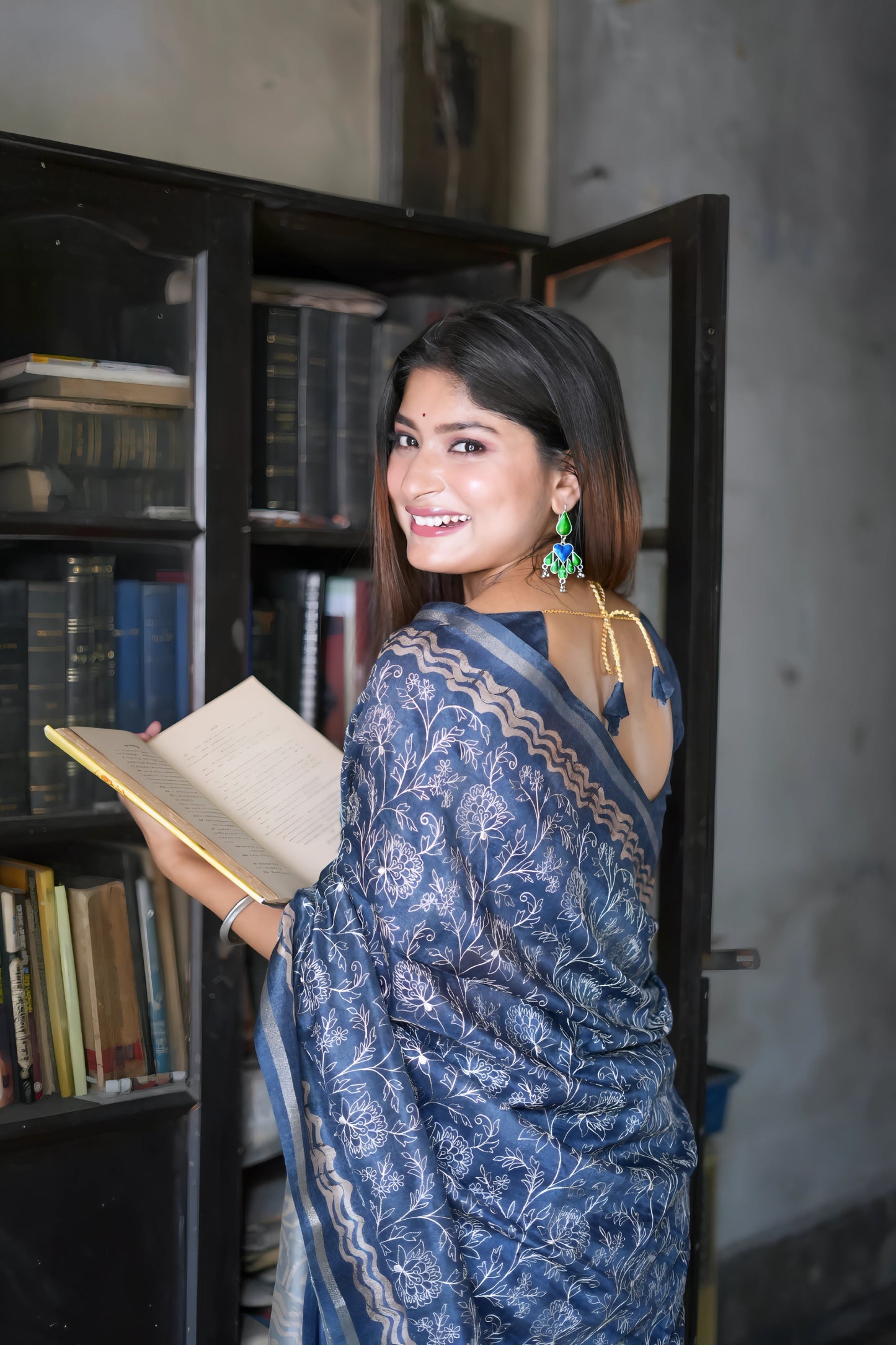 Woman wearing a blue floral printed saree with a matching blouse and statement earrings, standing beside a vintage black bookshelf filled with books and a green paneled door in a heritage-style room.