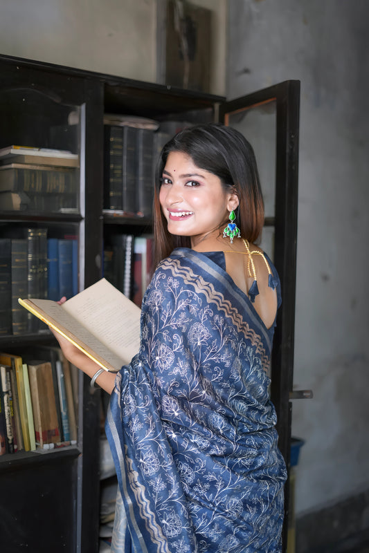 Woman wearing a blue floral printed saree with a matching blouse and statement earrings, standing beside a vintage black bookshelf filled with books and a green paneled door in a heritage-style room.