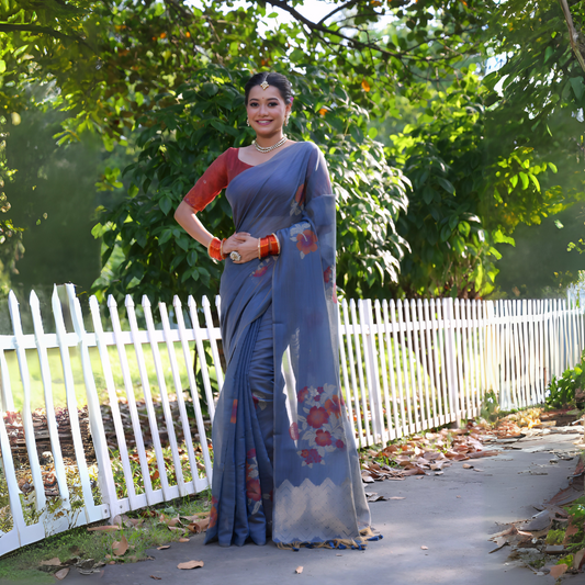Woman wearing a blue silk saree with red floral prints and a contrasting red blouse, accessorized with pearl jewelry and orange bangles, standing beside a white picket fence in a sunlit garden with lush green trees in the background.