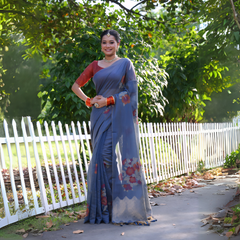 Woman wearing a blue silk saree with red floral prints and a contrasting red blouse, accessorized with pearl jewelry and orange bangles, standing beside a white picket fence in a sunlit garden with lush green trees in the background.