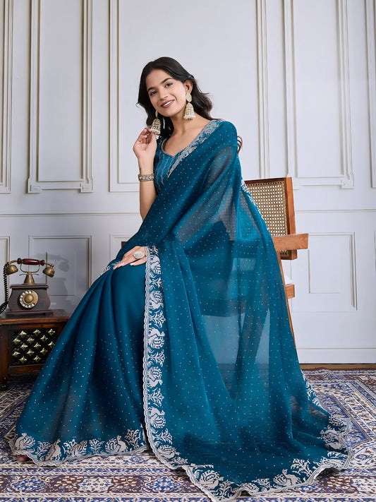Woman wearing a teal blue georgette saree embellished with sparkly sequins and an ornate silver embroidered floral border, paired with a matching blouse with delicate embroidery. She stands elegantly in a classic white paneled room, accessorized with statement jhumka earrings and bangles, next to a vintage rotary telephone on a carved wooden side table and a modern wooden armchair, all atop a richly patterned rug for a sophisticated, timeless look.