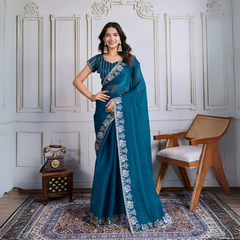 Woman wearing a teal blue georgette saree embellished with sparkly sequins and an ornate silver embroidered floral border, paired with a matching blouse with delicate embroidery. She stands elegantly in a classic white paneled room, accessorized with statement jhumka earrings and bangles, next to a vintage rotary telephone on a carved wooden side table and a modern wooden armchair, all atop a richly patterned rug for a sophisticated, timeless look.