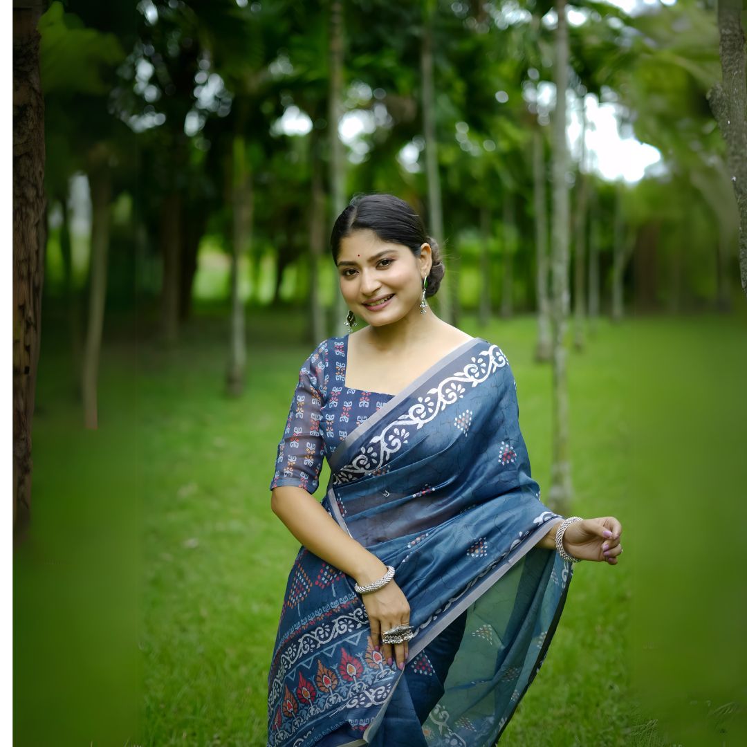 Woman wearing a blue handloom cotton saree with white tribal-inspired border and geometric patterns, paired with a matching printed blouse and silver jewelry, standing gracefully in a lush green garden with tall trees in the background.