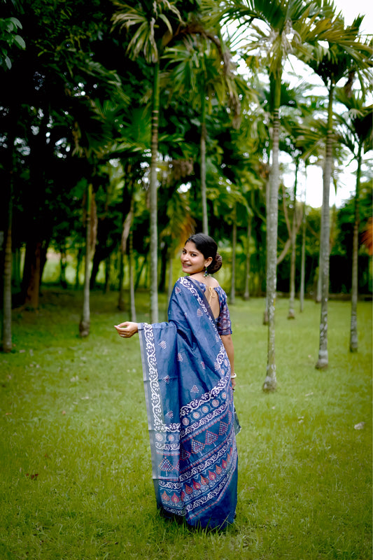 Woman wearing a blue handloom cotton saree with white tribal-inspired border and geometric patterns, paired with a matching printed blouse and silver jewelry, standing gracefully in a lush green garden with tall trees in the background.