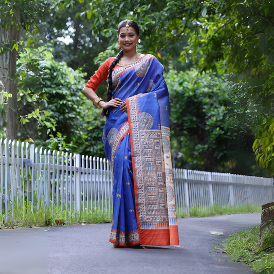  Woman wearing a blue Madhubani-printed saree with intricate traditional motifs and an orange blouse, accessorized with bangles and a long braid, standing confidently on a garden path lined with white fencing and lush green foliage in the background.
