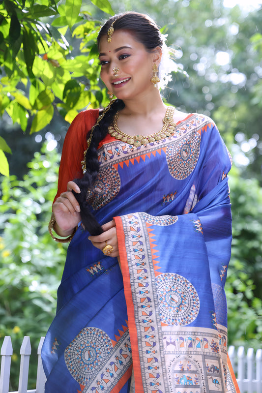  Woman wearing a blue Madhubani-printed saree with intricate traditional motifs and an orange blouse, accessorized with bangles and a long braid, standing confidently on a garden path lined with white fencing and lush green foliage in the background