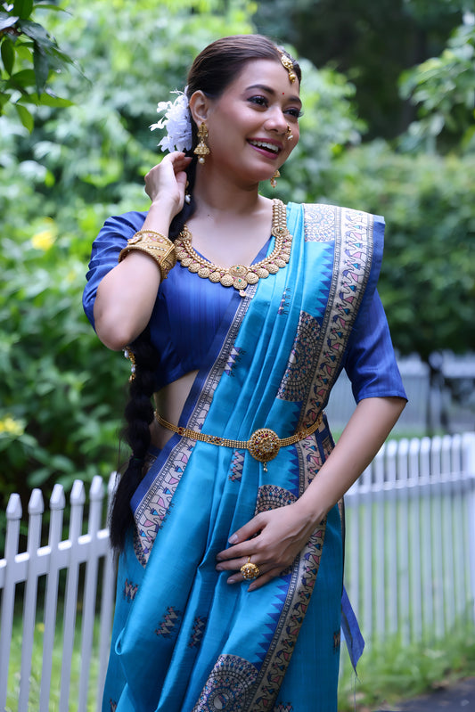 Woman wearing a blue Madhubani-printed saree with an intricate white border and traditional motifs, paired with a matching blue blouse and gold jewelry, standing gracefully on a garden path lined with a white picket fence and surrounded by lush green foliage.