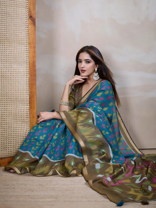  Woman wearing a blue silk saree featuring vibrant pink and olive green ikat-inspired abstract patterns, paired with a matching blouse and elegant gold bangles. She poses gracefully in a contemporary studio setting with a neutral textured wall, a tall woven cane screen, and rustic wooden flooring, highlighting the saree’s flowing pallu and artistic design.