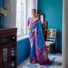Woman wearing a blue Patola silk saree with purple and gold borders, paired with a purple blouse and traditional jewelry, standing gracefully by a window in a vintage-inspired room with teal blue walls, wooden furniture, and floral decor.