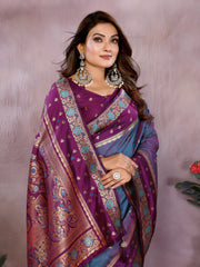 Woman wearing a blue and purple Banarasi silk saree with an ornate golden border and intricate traditional motifs, paired with a matching purple blouse adorned with gold detailing and statement earrings. She poses gracefully in a modern studio featuring potted green plants and blush-toned vases on a neutral backdrop.