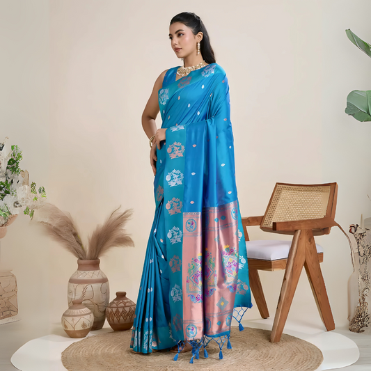 Woman wearing a vibrant blue silk saree with intricate pink and silver woven border and pallu, paired with a matching blouse and gold jewelry, elegantly posing beside a wooden chair in a minimalist studio setting with neutral decor, ceramic vases, and pampas grass.

Related
blue-silk-saree-pink-border-modern-studio
Woman in a vibrant blue silk saree with pink border and intricate details in a modern studio setting.